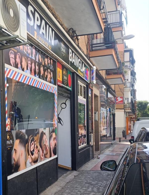 Fachada de Spain Bangla Peluquería en Villaverde Bajo: rótulo negro con letras blancas, escaparate con cenefa de barbero y el poste de rayas girando.